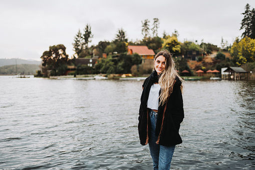 Young blonde warm woman standing on a wooden dock, in a beautiful lake on a cloudy afternoon. Copy space
