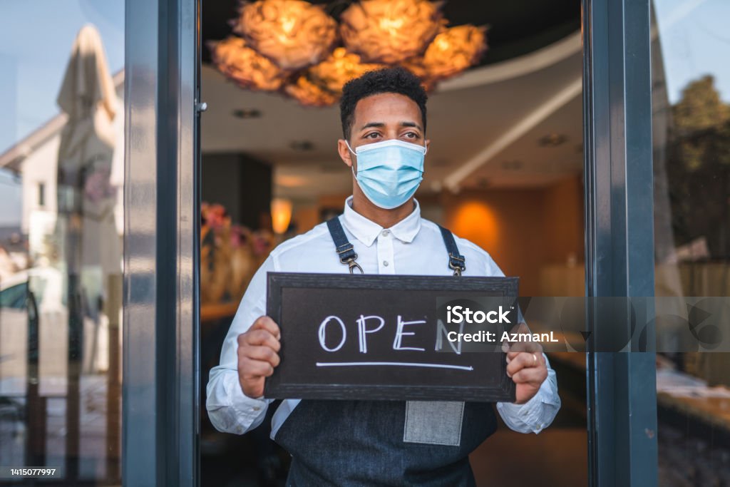 Mixed Race Waiter Holding an "Open" Sign at the Entry of a Cafe With a Face Mask On Open sign on an entry of a small classy cafe held by a mixed race mid adult waiter wearing a face mask. 35-39 Years Stock Photo