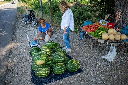 Ksamil, Albania July 4, 2022 A woman tourist buys vegetables from a woman at a  roadside farm stand selling tomatoes and watermelons.