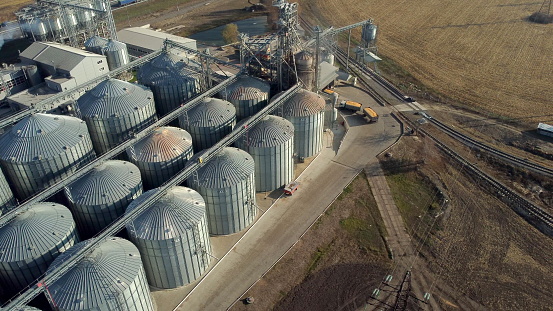Aerial Drone View Truck Full of Grain Rides Near Large Metal Silos of Grain Elevator on Sunny Day. Top View Metal Complex of Grain Elevator. Flight Over Elevator Tanks for Drying and Storage of Grain.