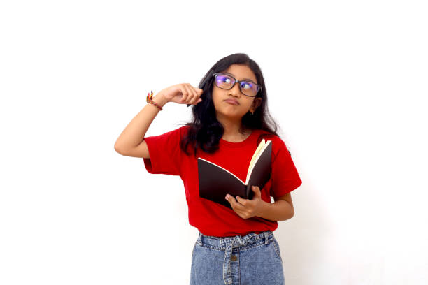 istock Thoughtful asian schoolgirl standing while holding a book and thinking an idea. Isolated on white. 1415110961