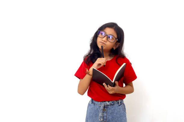 Thoughtful asian schoolgirl standing while holding a book and looking sideways. Isolated on white