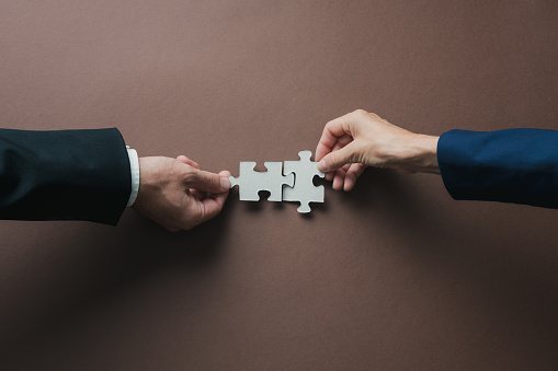 Teamwork and partnership conceptual image - hands of a businessman and businesswoman joining blank matching puzzle pieces over brown background.