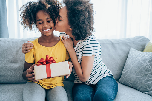 Shot of a loving mother giving her daughter a gift. Mother surprise her little girl with present while she is sitting on the sofa in the living room at home.