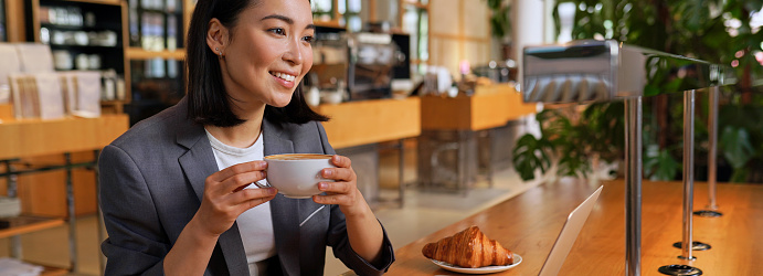 Happy smiling young Asian business woman entrepreneur or student having coffee break relaxing after remote work or learning, working on laptop computer sitting at table in coffeeshop. Banner