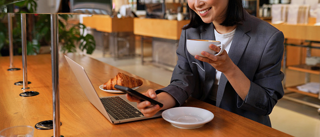 Young Asian business woman wearing suit drinking coffee using smartphone in cafe. Happy smiling female professional working holding mobile phone using smartphone texting messages on cellphone.