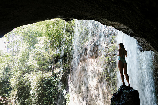 Woman in bikini sit on rock under falling water of Suwat waterfall in tropical jungle. Nature day tour, hiking activity adventure and fun at family tourist camp on summer vacation in Bali island
