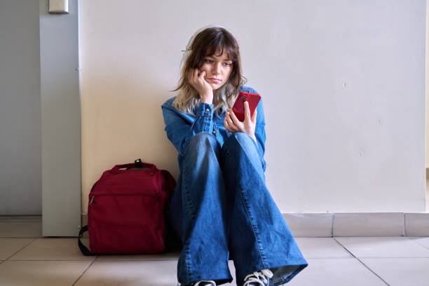istock Sad young female student with backpack smartphone sitting on the floor 1415127199