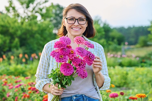 Portrait of middle aged woman with bouquet of flowers outdoor. Smiling happy 40s female looking at camera, holding zinnia flowers, on summer day. Nature, beauty, maturity, people concept