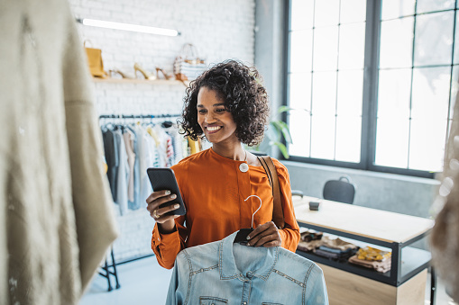 Young woman shopping clothes in thrift store. She is using smart phone while choosing clothing.