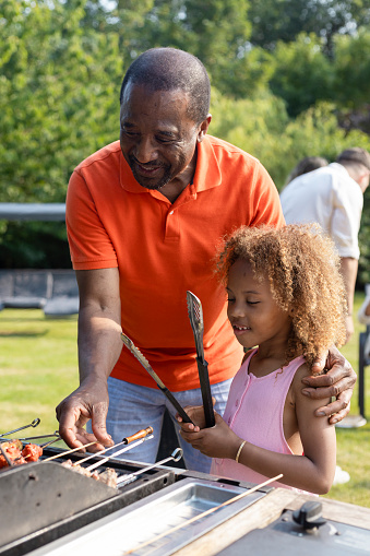 A senior grandfather preparing a barbecue in his back garden on a bright summers day, he is teaching his young granddaughter how to cook on the barbecue, all the meat on the barbecue is halal.