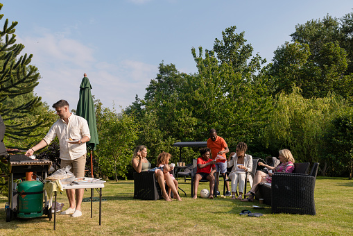 A wide-view shot of a multi-gen family barbecue, they are enjoying food and drinks in a back garden on a bright summers day, all the meat on the barbecue is halal.