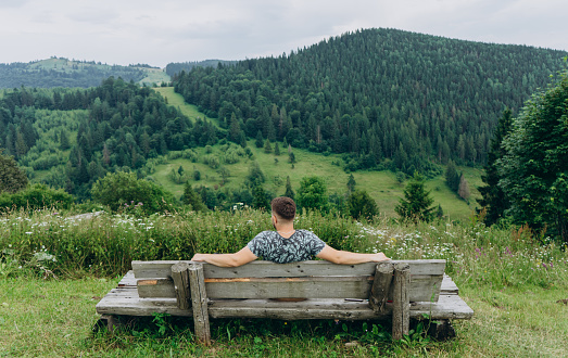 A man in a T-shirt sits with his back to the photographer resting after climbing the mountains. Young man sitting on a wooden bench on a meadow on a sunny hot day.