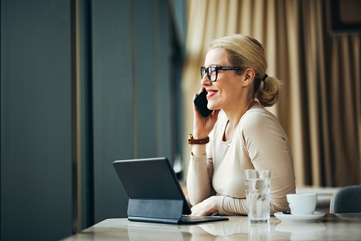 Happy business woman speaking on the phone while sitting at restaurant desk with her digital tablet, cup of coffee and glass of water.
