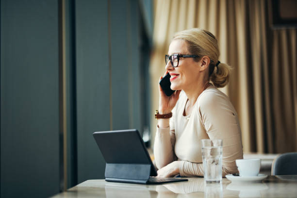 Smiling Businesswoman Talking On A Mobile Phone In The Cafe Happy business woman speaking on the phone while sitting at restaurant desk with her digital tablet, cup of coffee and glass of water. older blonde woman stock pictures, royalty-free photos & images