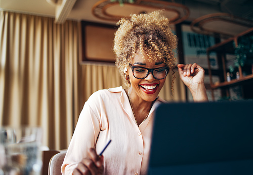 Cheerful smiling African-American business woman teleconferencing using a wireless earphones and a tablet while sitting at restaurant desk.
