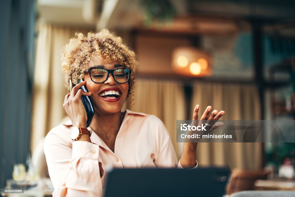 Smiling Businesswoman Talking On A Mobile Phone In The Cafe Happy African-American business woman speaking on the phone while sitting at restaurant desk with her digital tablet. Using Phone Stock Photo