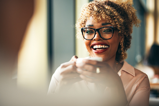 Smiling African-American businesswoman drinking cup of coffee and talking with unrecognizable client or coworker while sitting together in the restaurant.