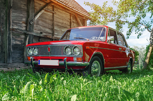 Close-up view of red vintage Lada car in countryside. Old wooden barn background. Selective focus of green grass lit by sunlight.