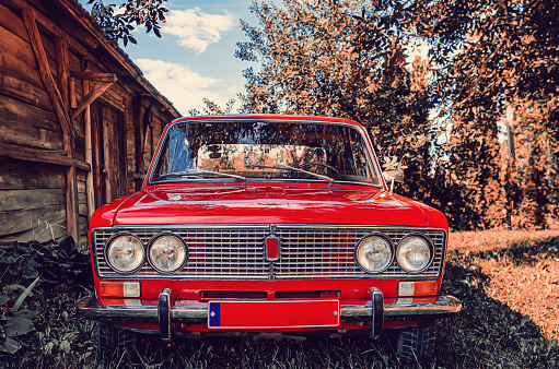 Front view of red vintage Lada car in countryside. Old wooden barn from side. Old red Soviet car.