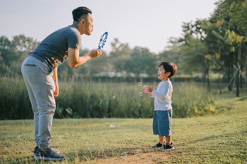 Asian chinese father playing with son at park blowing bubbles spending bonding time together