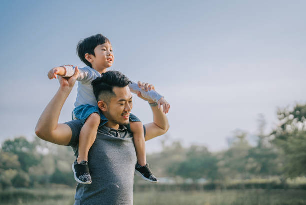 asian chinese father carrying his son on shoulder at public park enjoying bonding time together  during weekend leisure time stock photo