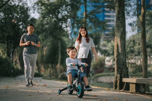 Asian Chinese boy learning cycling in public park with his 2 parents during weekend morning