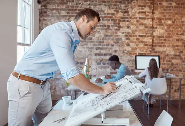 Photo of Architect, design engineer or building contractor drawing a plan on a drafting table for a project or development in his office. A male designer doing a blueprint sketch at an architecture company