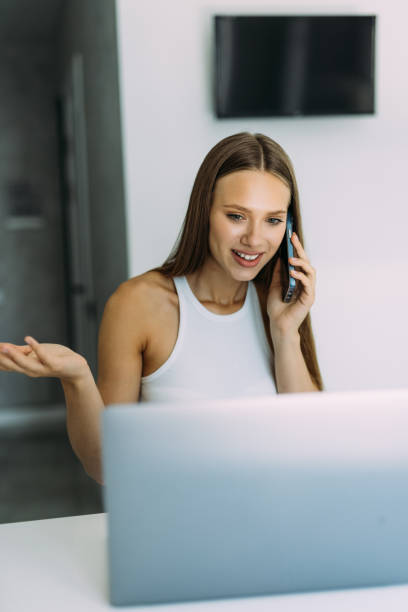istock Young woman sits at the kitchen table using a laptop and talking on a ce phone 1415155929