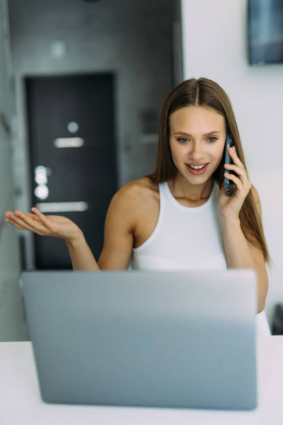 istock Young cheerful happy woman indoors at home at the kitchen using laptop computer talking by mobile phone. 1415156343