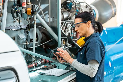 Female aviation mechanic referring to helicopter engine repair checklist in digital tablet, side view. Engineer in protective workwear fixing air vehicle motor in hangar, medium shot