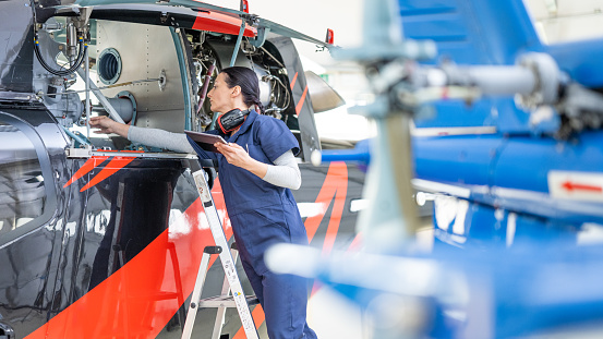 Female helicopter mechanic in uniform standing on ladder and looking into jet medium shot. Engineer working in air helicopter hangar, front side view