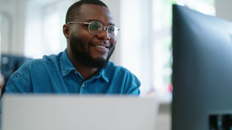 African businessman using laptop and looking at computer monitor sitting at desk