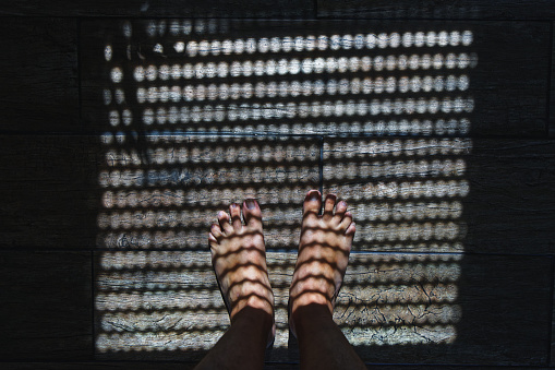 top view of feet in shade of roller blind casting wavy pattern on wooden tiles floor