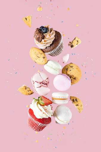 Colorful decorated Broken chocolate chip cookies, cupcakes and macaroons falling in motion on pink background with sprinkling and pieces with crumbs. Sweet and various pastries flying.