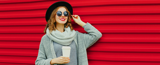 Portrait of beautiful young woman with cup of coffee wearing black round hat, gray knitted cardigan sweater on red background