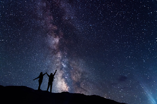 Two hiker silhouette stands on the hill and looking at the bright milky way galaxy in the starry night.