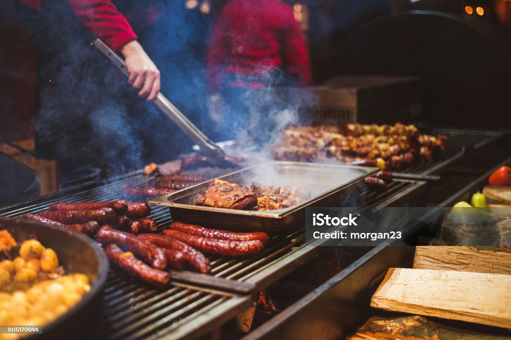 Outdoor street food festival. Chief cooking sausages, meat and potatoes cooking on an outdoor griddle Barbecue Grill Stock Photo