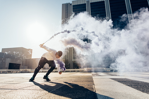 Sporty man practicing parkour and free running by doing a flip with a smoke grenade. Free runner in action over urban city background.
