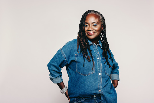 Fashionable mature woman looking at the camera while standing against grey background. Stylish woman with dreadlocks wearing a denim jacket and make-up in a studio.