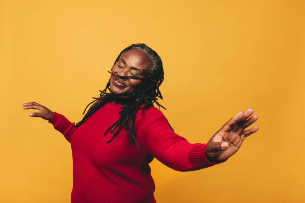 Joyful black woman dancing in a studio Joyful mature woman dancing and having fun while standing against a yellow background. Happy black woman with dreadlocks embracing her natural hair with pride. danser stock pictures, royalty-free photos & images