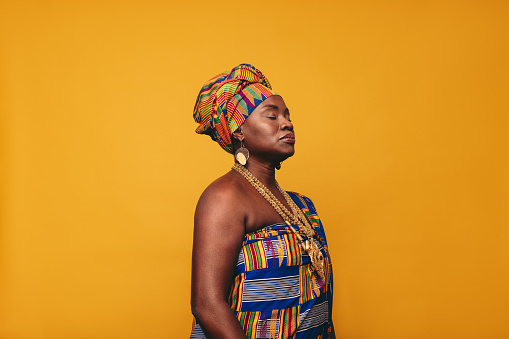 Mature woman wearing an African traditional attire against a yellow background. Confident black woman dressed in colourful Kente cloth and golden jewellery. Woman embracing her rich West African culture.