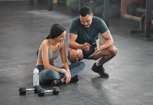 Personal trainer, coach or fitness instructor helping an active and fit woman in the gym. Young female athlete sitting down and managing her workout routine or schedule with her exercising partner