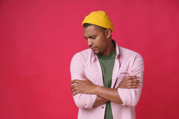 istock Serious passionate handsome African American young man posing with hands folded and head nodede sideways down wearing casual pink shirt and yellow hat smiling on camera isolated on red background 1415177380