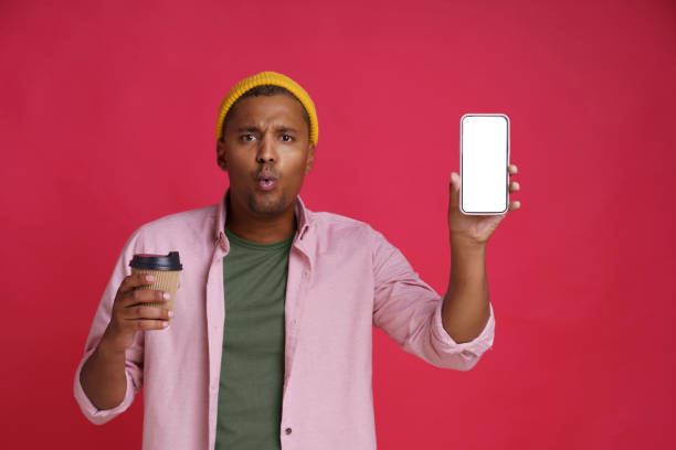 istock Handsome young African American man, guy using smartphone while drink coffee holding in hands with oooh excitement on his face showing smartphone with white screen. Mobile app advertisement 1415177383