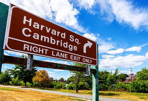 Boston, Massachusett, USA - August 14, 2022: Close-up of a Storrow Dirve highway exit sign along the Charles River pointing the way to Harvard Square in Cambridge. Storrow Drive runs along the Boston riverbank of the Charles River. In the distance is the Eliot House building of Harvard University's Cambridge campus.