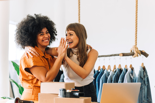 Cheerful online store owners embracing each other in their thrift store. Two happy businesswomen celebrating their success as a team. Female entrepreneurs running an e-commerce small business.