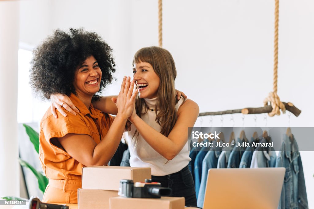 Cheerful online store owners high-fiving each other in their thrift store Cheerful online store owners embracing each other in their thrift store. Two happy businesswomen celebrating their success as a team. Female entrepreneurs running an e-commerce small business. Happiness Stock Photo