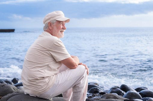 Senior man with hat sitting on the beach relaxing enjoying retirement. Elderly bearded male enjoying sunset at sea, horizon over water