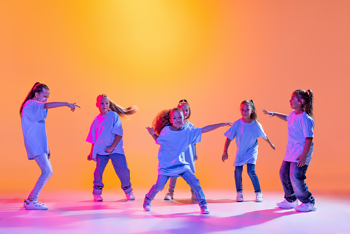 Happy children dancing. Group of children, little girls in sportive casual style clothes dancing in choreography class isolated on orange background in purple neon light. Concept of music, fashion, art